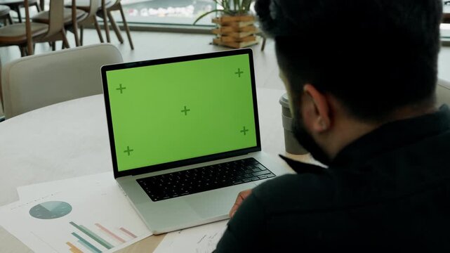 Indian man analyses commercial data with diagrams at laptop with green chromakey screen in office rear view. Marketer checks market trends to build strategy at corporate workplace