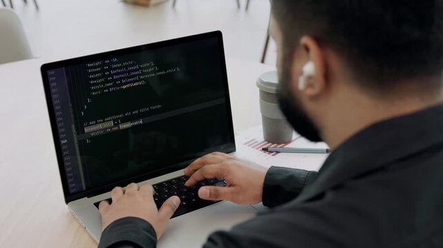 Indian man programmer with earphones writes code on laptop as sits at table closeup. Skilled software engineer develops app with portable computer backside view. Computing science