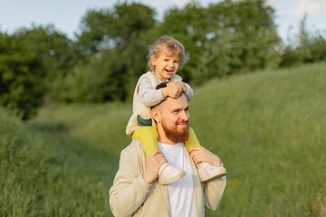 Father with a red beard carrying his toddler on his shoulders while walking outdoors on a sunny day. Warm natural light, joyful family moment and peaceful countryside atmosphere.