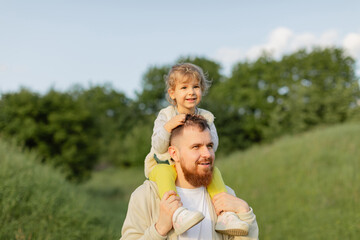 Father with a red beard carrying his toddler on his shoulders while walking outdoors on a sunny day. Warm natural light, joyful family moment and peaceful countryside atmosphere.