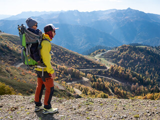 A man and his child in backpack carrier are hiking and looking at autumn mountains. Rosa Khutor resort in Krasnaya Polyana (Sochi) at sunny day. Not AI