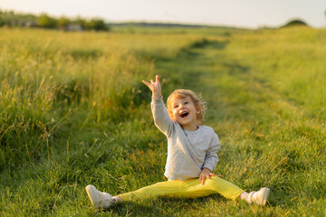 Happy toddler sitting on the grass at golden hour, laughing and reaching up with joy. Candid childhood moment in a sunny outdoor meadow.
