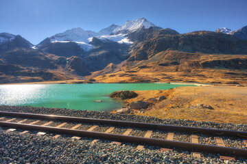 Obraz premium Scenic railway tracks running along turquoise lake Bianco with snowy alpine peaks on autumn sunny day. Swiss Alps landscape near Bernina Pass, train route in Switzerland. Railway station. Travel