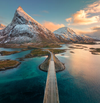 Aerial view of the Fredvang bridge and coastal road at sunset. Lofoten Islands, Norway. Turquoise sea, rocky islands, snowy mountains in warm evening light. Nordic landscape and Arctic road. Top view