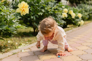 Barefoot toddler drawing with chalk on a garden pathway surrounded by blooming yellow peonies. Natural light, playful creativity, summer childhood moment.