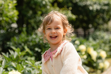 Smiling toddler enjoying a sunny day in the garden, surrounded by green foliage and soft yellow flowers. Bright natural light and joyful emotion in an outdoor childhood moment.