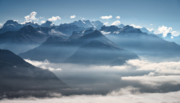 Mountains above clouds in the Swiss Alps with snow-covered peaks and blue haze at sunset. Aerial view with evening light, fog in valleys, clear sky over rugged alpine terrain. Top view. Switzerland