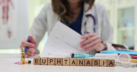 Wooden blocks spell word Euthanasia beside syringe vial and pills on medical desk. Woman doctor writes notes evaluating euthanasia ethics case