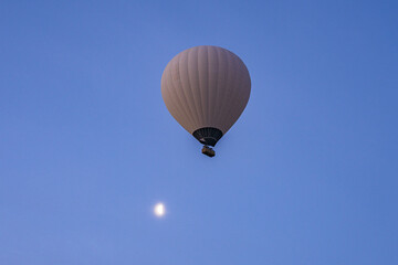 View of a solitary hot air balloon ascends into the tranquil, gradient blue sky, with a faint moon adding a touch of magic, Love valley, Cappadocia, Turkey.