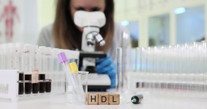 Abbreviation HDL appears on wooden blocks near microscope and test tubes. Woman lab scientist studies lipid profile noting high density fraction
