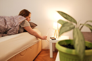 A woman reaches from bed to switch on a bedside lamp, creating a warm and cozy morning atmosphere in a simple home interior.