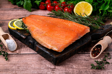 Hot smoked trout fillet on a wooden board. Lettuce, cherry tomatoes, lemon, rosemary and pepper are arranged around