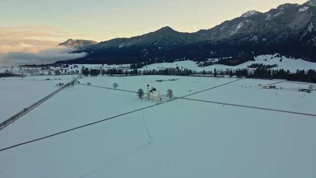 Aerial view of a church surrounded by a vast snow-covered field, roads, and mountains in the distance, Schwangau, Bavaria, Germany.