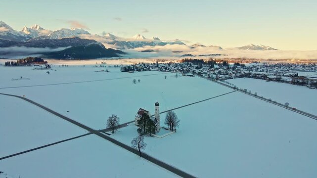 Aerial view of a church surrounded by a vast snowy field, with the village nestled against the backdrop of majestic mountains, Schwangau, Bavaria, Germany.