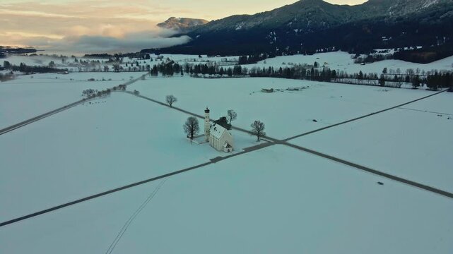 Aerial view of a lone church surrounded by stark white snowfields and dark roads under the winter sky, Schwangau, Bavaria, Germany.