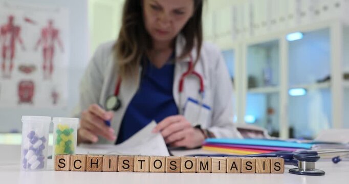 Wooden blocks spell word Schistosomiasis near sample boxes and pills. Woman infection specialist fills patient form studying tropical worm disease