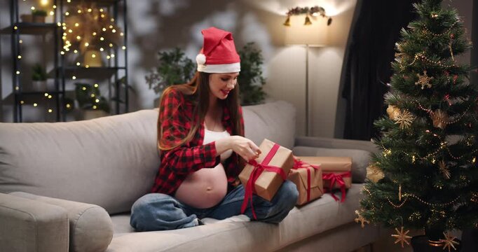 A pregnant woman wearing a Santa hat opens a Christmas present on a couch, surrounded by festive decorations. A pregnant woman in a Santa hat looks at sonogram images near a Christmas tree