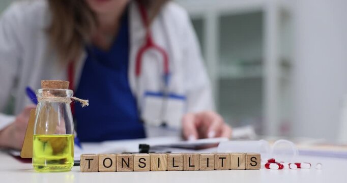 Wooden blocks spell word Tonsillitis near bottle with liquid and stethoscope on table. Woman clinician writes throat infection notes adjusting therapy