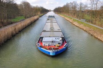 A barge sails through a calm canal in Germany. Loaded or empty, it is clearly on its way, symbolizing movement and logistics on Europe&rsquo;s waterways.	