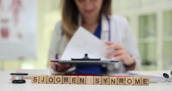 Wooden blocks spell words Sjogren Syndrome while woman doctor holds clipboard. Woman rheumatologist writes notes about autoimmune dryness problem