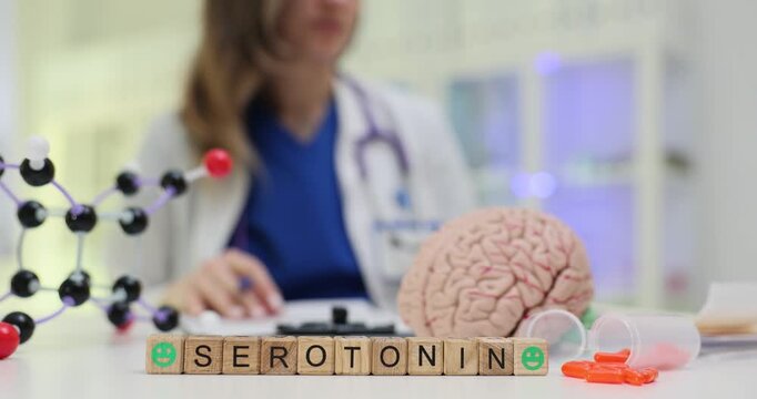Wooden blocks spell word Serotonin near brain model and capsules on desk against doctor. Woman neurologist writes notes monitoring serotonin level