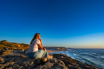 Beautiful middle-aged woman sitting on cliff overlooking Monte Clerigo beach and ocean on summer evening. Algarve coast in Portugal. Side view