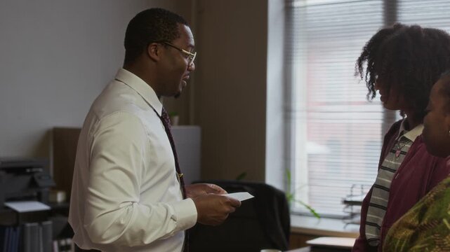 Medium shot of male school principal handing envelope to mother and her teenage daughter, they opening it together and reacting with happiness in office