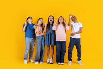 Fototapeta premium Preteen kids standing together pointing upward on yellow studio background. Curious children showing interest, imagination and a positive school lifestyle moment