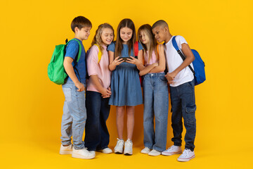 Obraz premium Group of school kids gathered around girl with smartphone in hands, standing on bright studio background. Friendly children sharing modern digital moment together