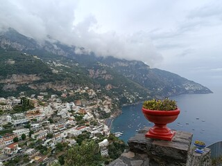 A serene Positano panorama showing clifftop homes, lush hills, and boats scattered along the turquoise coast, framed by soft clouds drifting over the Amalfi landscape.
