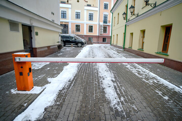 Closed orange boom gate guards the entrance to snow-dusted courtyard lined with classic buildings. Lowered security gate blocks a narrow cobblestone path. Automatic barrier arm