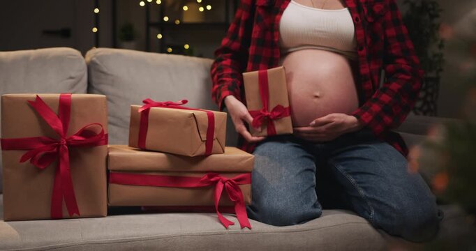 A pregnant woman sits on a couch holding a gift, surrounded by wrapped presents, anticipating the holiday season. A smiling pregnant woman wearing a Santa hat opens a present on Christmas day