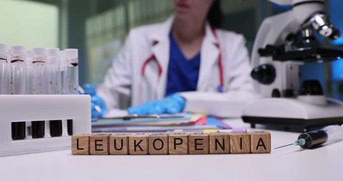Word Leukopenia on wooden cubes near blood samples in medical laboratory. Female doctor researches decrease in number of leukocytes with microscope