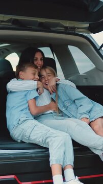 Charming loving joyful mom hugging her cheerful teen son and daughter while they together sitting in auto trunk,stopped in the green field during travel countryside,family concept