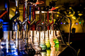A row of colorful hookahs with coiled hoses and glowing bases on a reflective surface. Warm lighting and decor create a cozy lounge atmosphere.