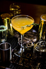 A yellow-orange cocktail served in a coupe glass with rosemary garnish. Bar tools and lighting create a refined mixology ambiance.