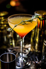 A cocktail with gradient colors from red to yellow-orange served in a stemmed glass with rosemary garnish. Reflections and tools enhance appeal.