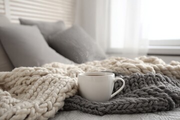 White mug with hot drink sits on chunky gray and cream knit blankets, comfy bedroom scene.