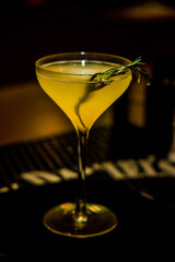 A yellow cocktail in a stemmed glass with rosemary garnish, set in a dimly lit bar setting.