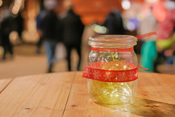 Glass jar with warm LED string lights and decorative red ribbon placed on wooden tabletop, with blurred crowd movement and illuminated stalls in evening market background