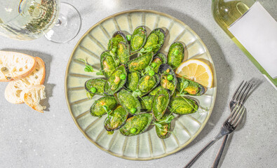 Gourmet baked mussels in green herb sauce served with lemon slices, white wine and bread on a light stone background, top view