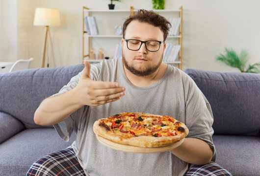 Overweight man resists diet holding pizza. He inhales aroma on a sofa, hinting at obesity, cravings, and fastfood temptation during a quiet evening. Choose health over pizza tonight.