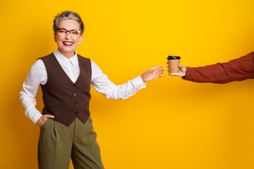 A confident mature woman in formalwear smiles as she receives a cup from a colleague against a bright yellow background showing collaboration and friendly teamwork