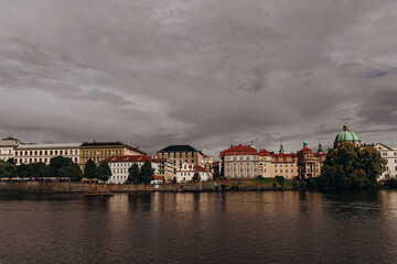 Obraz premium Historic waterfront architecture in Prague under cloudy skies reflecting on the river.