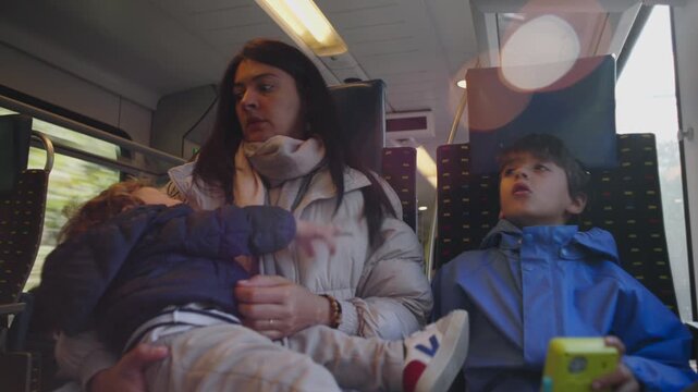 Mother sits on train holding restless toddler while older child looks upward creating authentic moment of overwhelmed parenting family travel fatigue and real everyday commute inside public transport