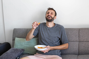 Smiling man sitting on a sofa and eating chips at home. Relaxed leisure time, enjoying a snack and laughing in a cozy living room environment. Casual lifestyle, comfort and positive mood indoors.