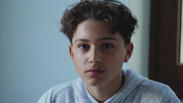 Teen boy looking straight into camera with calm serious expression inside home, creating intimate portrait moment with soft window backdrop and quiet emotional tone