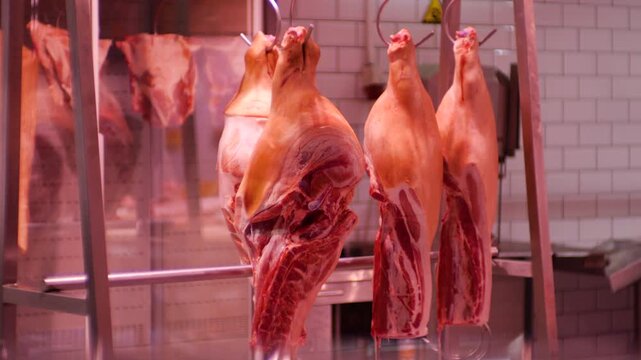 Raw pork carcasses hang from hooks in a butcher shop display cooler. The fresh meat is ready for sale to customers.