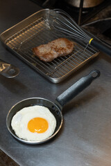 Fried egg cooking in a small pan on a stainless steel counter in a professional kitchen, with a piece of grilled meat resting on a wire rack in the background.