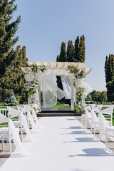 Elegant outdoor wedding ceremony setup with floral decorated pergola and white chairs.
